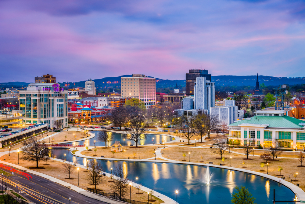 Evening cityscape with a winding water feature, trees, and lit buildings around a park in dusk light.